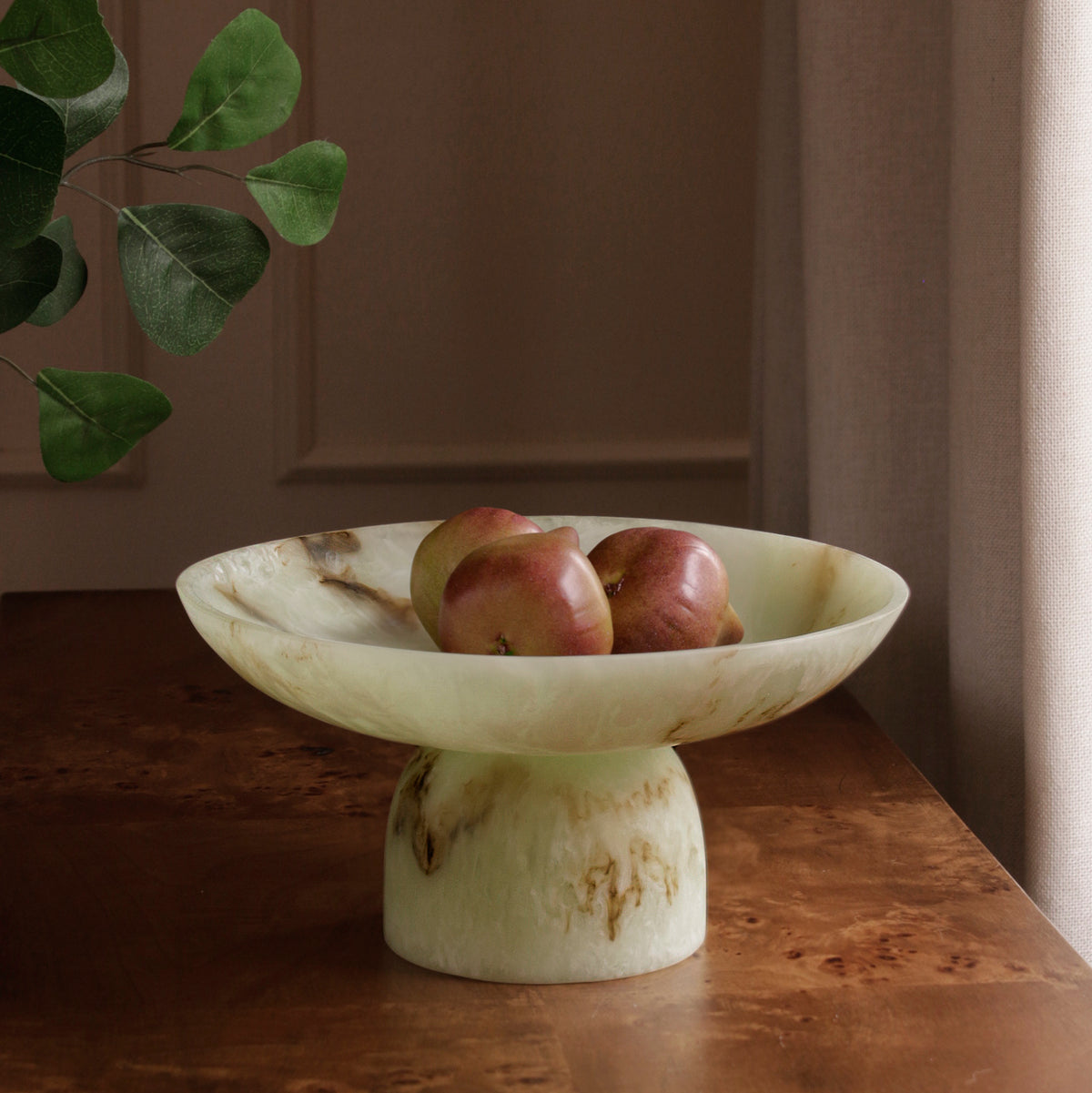 Jade swirl marble pattern resin pedestal bowl with apples on a wooden surface, with a plant in the background.