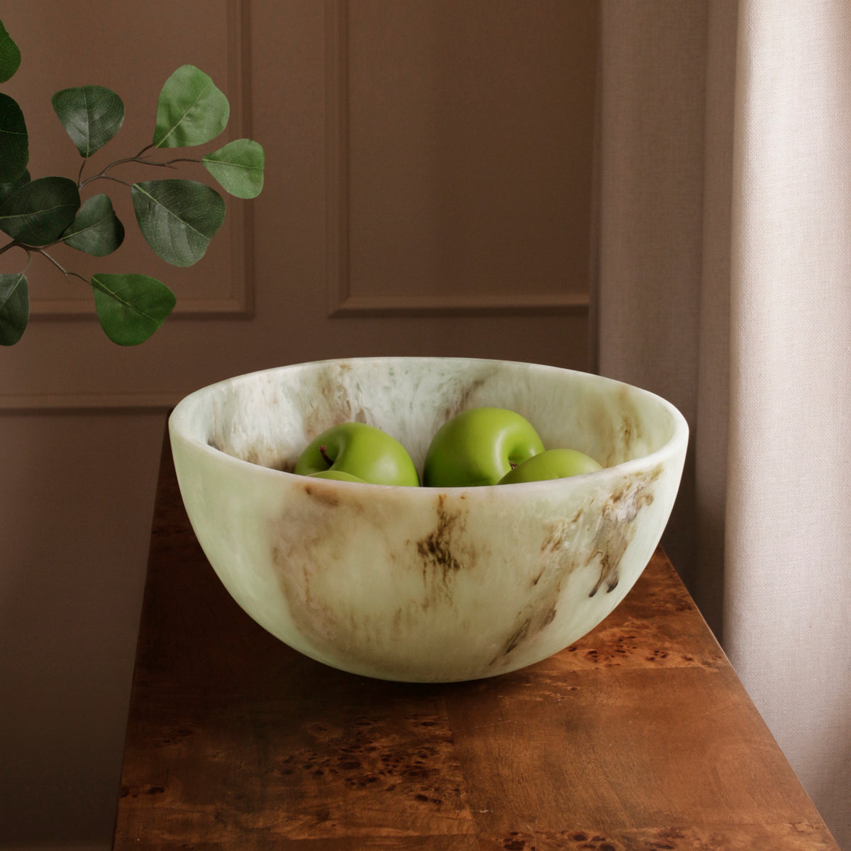 Jade swirl marble pattern resin bowl with green apples on a wooden table, with a plant in the background.
