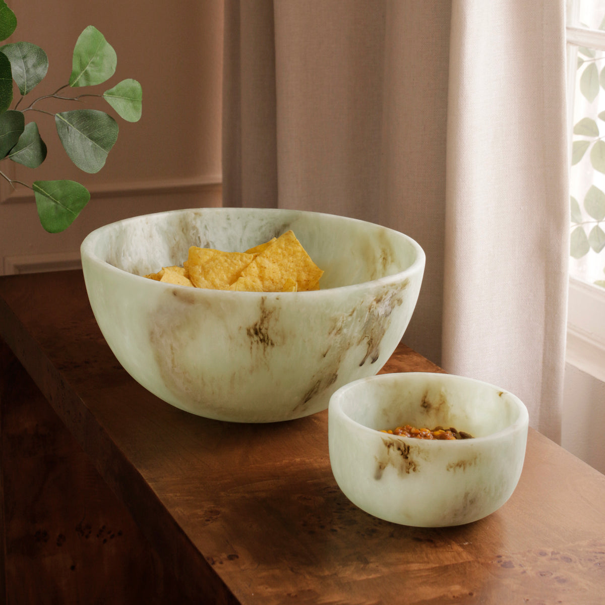Two Jade swirl marble pattern resin bowls of different sizes on a wooden surface with a plant in the background.