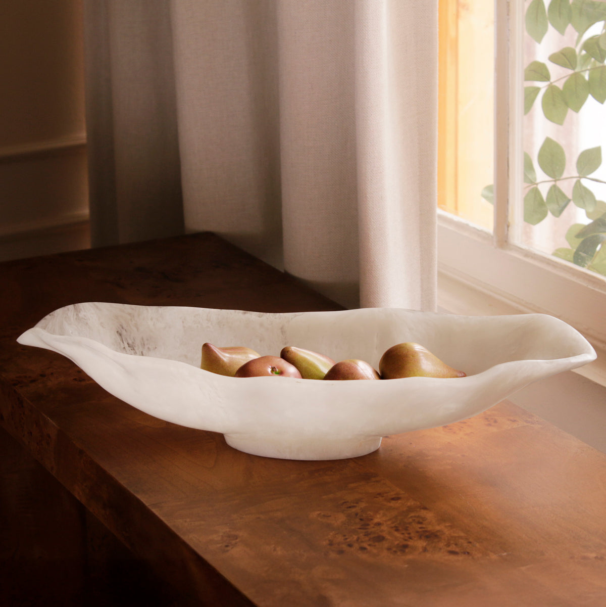 White resin bowl with fruits on a wooden table near a window with curtains.