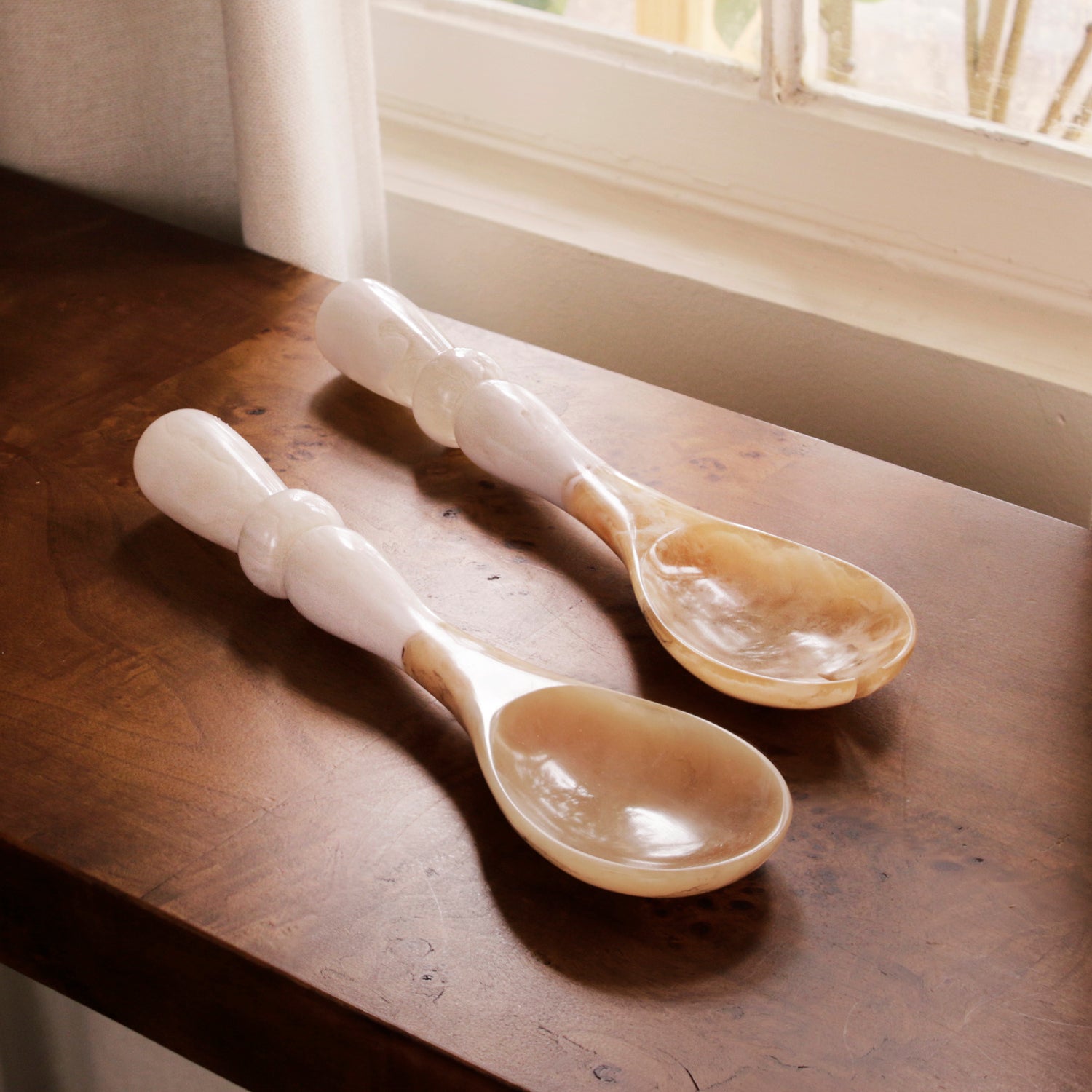 Two white resin handled spoons on a wooden surface with natural light coming through a window.