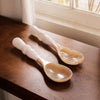 Two white resin handled spoons on a wooden surface with natural light coming through a window.