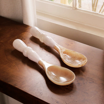 Two white resin handled spoons on a wooden surface with natural light coming through a window.